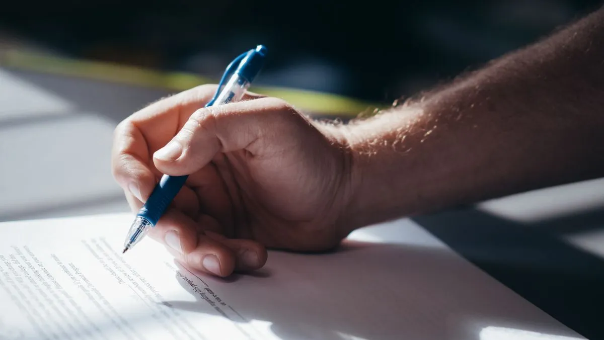 Hand signing compliance documentation with a blue pen on a desk